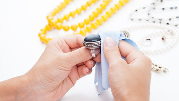 Caucasian hands using a blue polishing cloth to polich a large silver ring with a big hematite stone. Yellow beads and another beaded necklace in the background. The background is white. Kbeau jewelry guide to caring for your sterling silver jewelry.