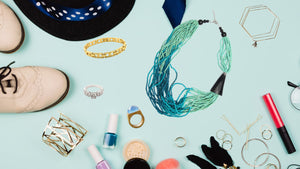 A flatlay photo with a light blue background with an assortment of jewelry laying on it. Also, a hat, shoes and lipstick are lying amongst the jewelry