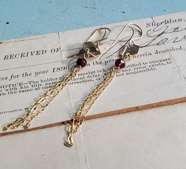 A pair of earrings lying on an old document from the 1800s. The earrings are a small shiny brass hexagon on ear wires. Below the hexagon is a wire wrapped garnet and hanging from the garnet are three chains. Chains and ear wire are 14 karat gold fill.